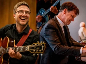 Een gitarist met bril speelt en lacht terwijl een pianist in pak geconcentreerd piano speelt tijdens een jazzoptreden.