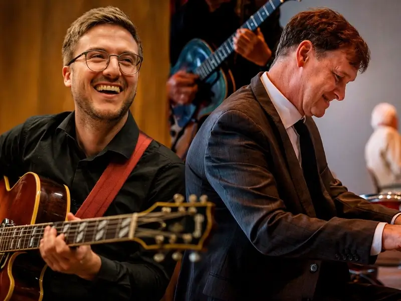 Een gitarist met bril speelt en lacht terwijl een pianist in pak geconcentreerd piano speelt tijdens een jazzoptreden.