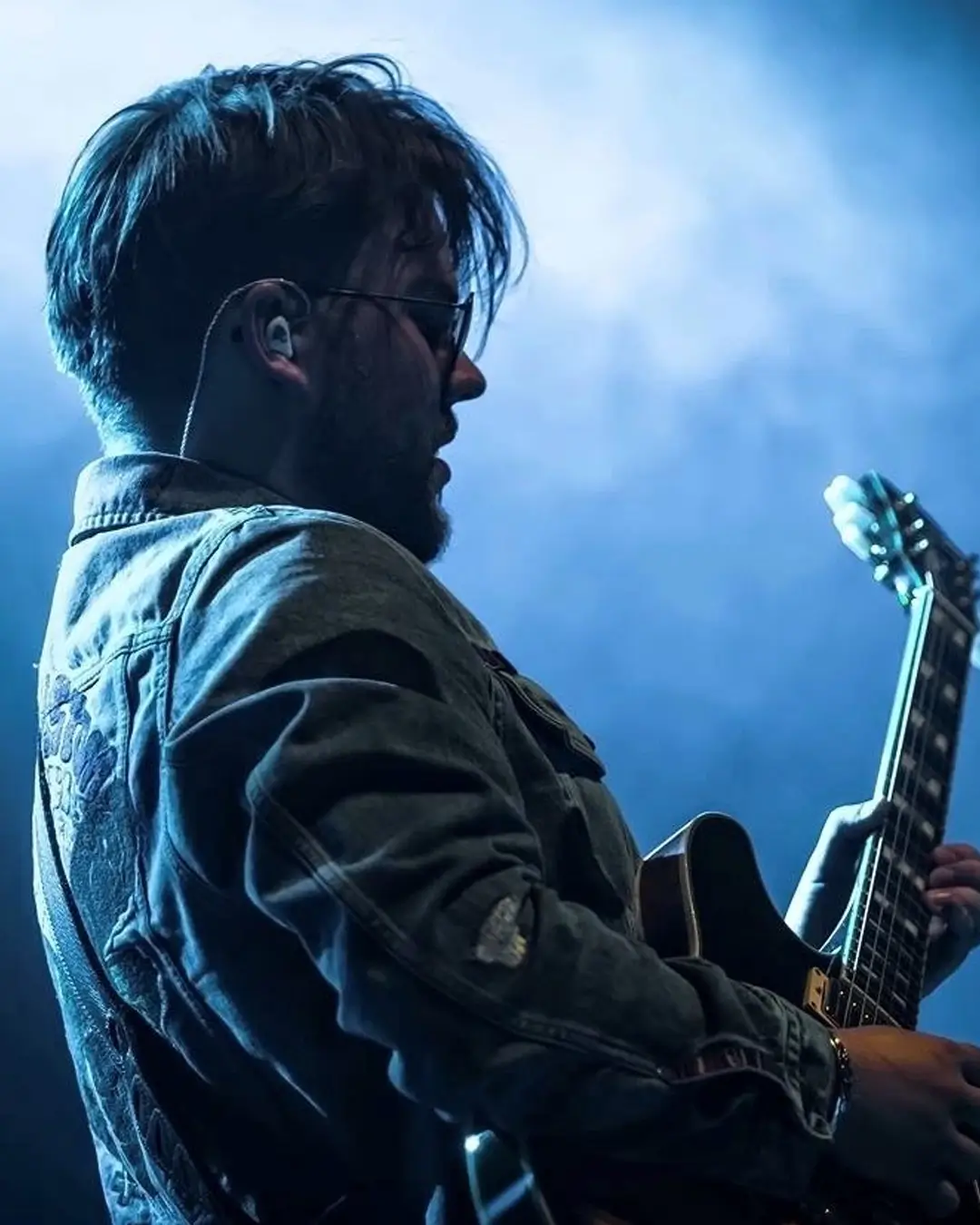 Zijaanzicht van Lorijn von Piekartz op het podium, spelend op een elektrische gitaar onder blauw podiumlicht en lichte rook, met in-ear monitors en een denim jas.
