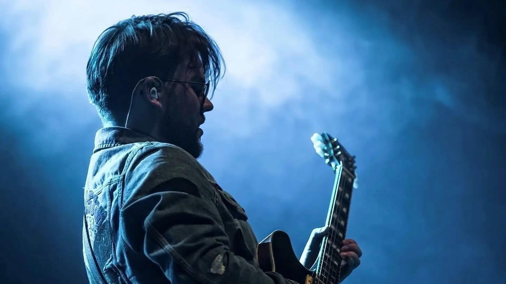 Zijaanzicht van Lorijn von Piekartz op het podium, spelend op een elektrische gitaar onder blauw podiumlicht en lichte rook, met in-ear monitors en een denim jas.
