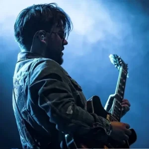 Zijaanzicht van Lorijn von Piekartz op het podium, spelend op een elektrische gitaar onder blauw podiumlicht en lichte rook, met in-ear monitors en een denim jas.