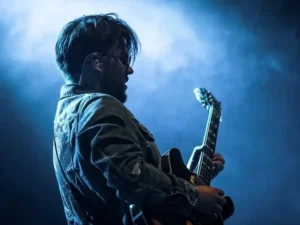 Zijaanzicht van Lorijn von Piekartz op het podium, spelend op een elektrische gitaar onder blauw podiumlicht en lichte rook, met in-ear monitors en een denim jas.