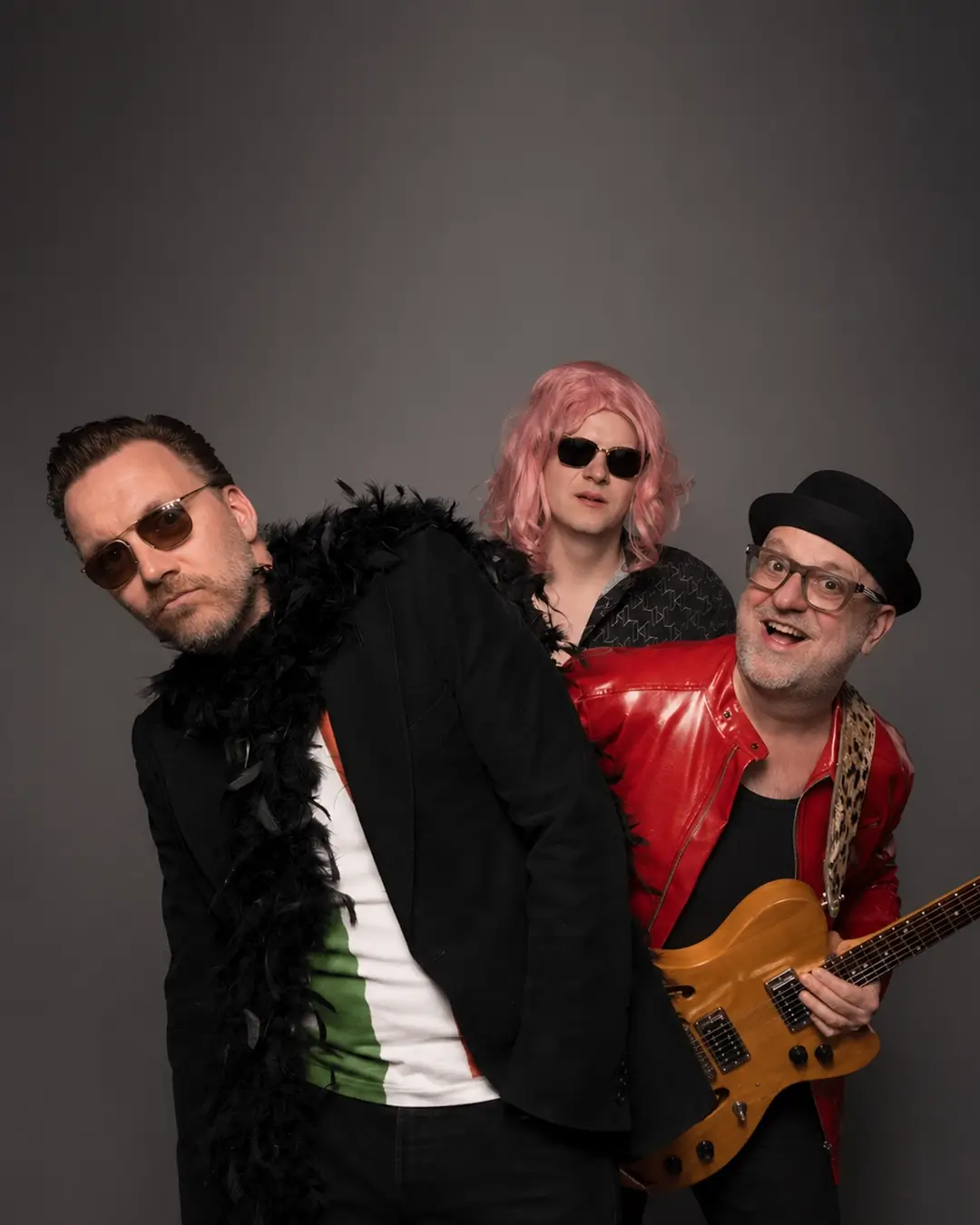 Portret van drie bandleden van Maison Prettyboy in een speelse pose: één met zonnebril en boa, één met roze pruik en zonnebril, en één met gitaar en rode jas, tegen een neutrale achtergrond.