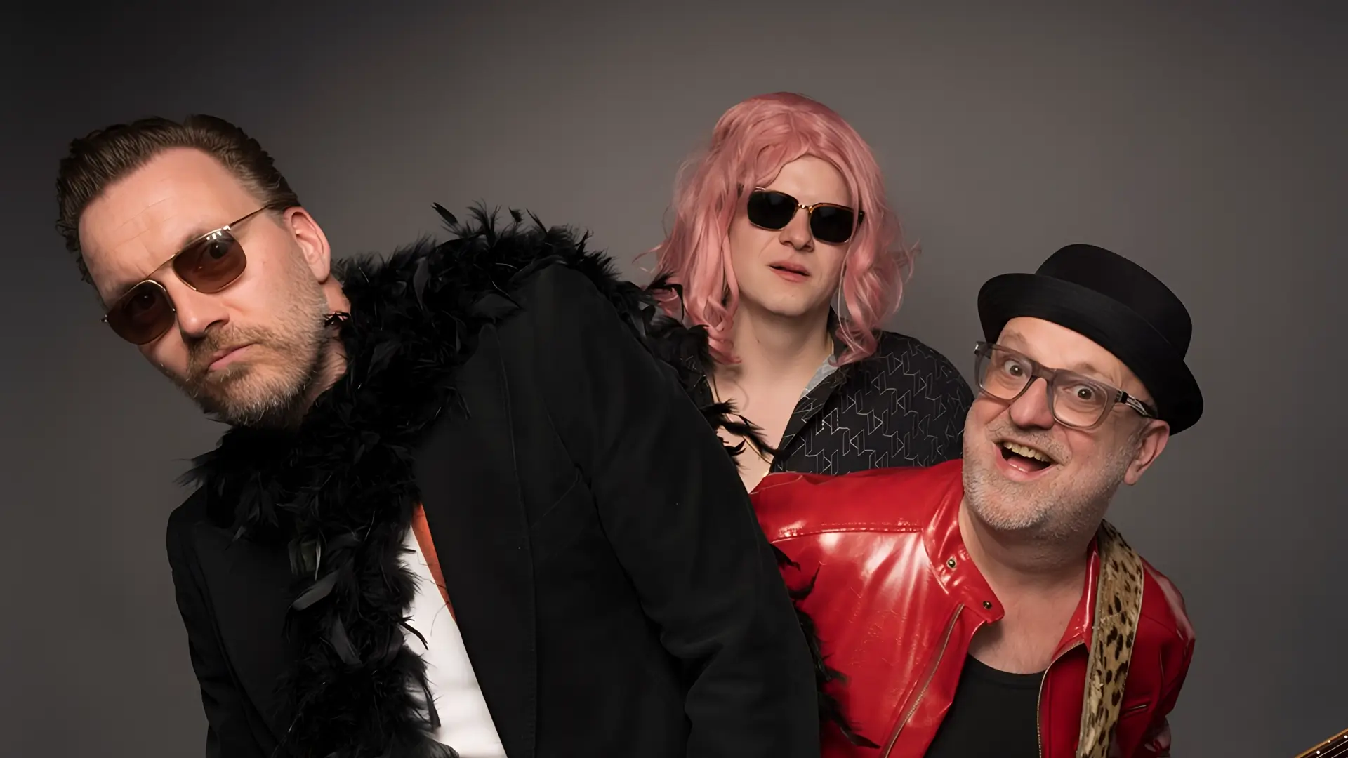Portret van drie bandleden van Maison Prettyboy in een speelse pose: één met zonnebril en boa, één met roze pruik en zonnebril, en één met gitaar en rode jas, tegen een neutrale achtergrond.