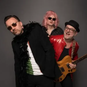 Portret van drie bandleden van Maison Prettyboy in een speelse pose: één met zonnebril en boa, één met roze pruik en zonnebril, en één met gitaar en rode jas, tegen een neutrale achtergrond.