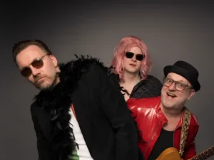 Portret van drie bandleden van Maison Prettyboy in een speelse pose: één met zonnebril en boa, één met roze pruik en zonnebril, en één met gitaar en rode jas, tegen een neutrale achtergrond.