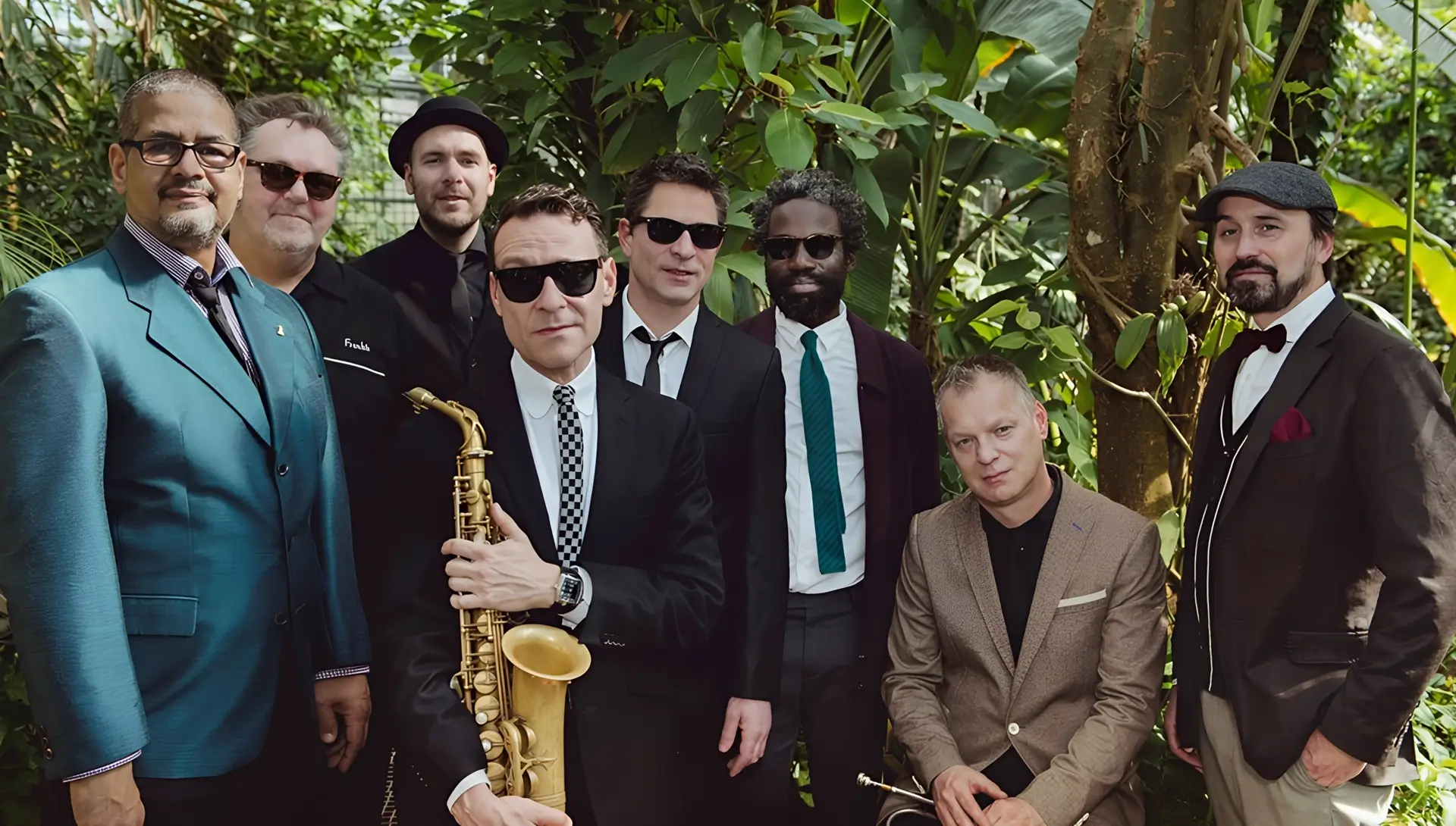 Groepsfoto van een jazzband in een groene, tropische omgeving; meerdere mannen in nette kleding en zonnebrillen poseren samen, met een saxofonist prominent vooraan die zijn instrument vasthoudt.