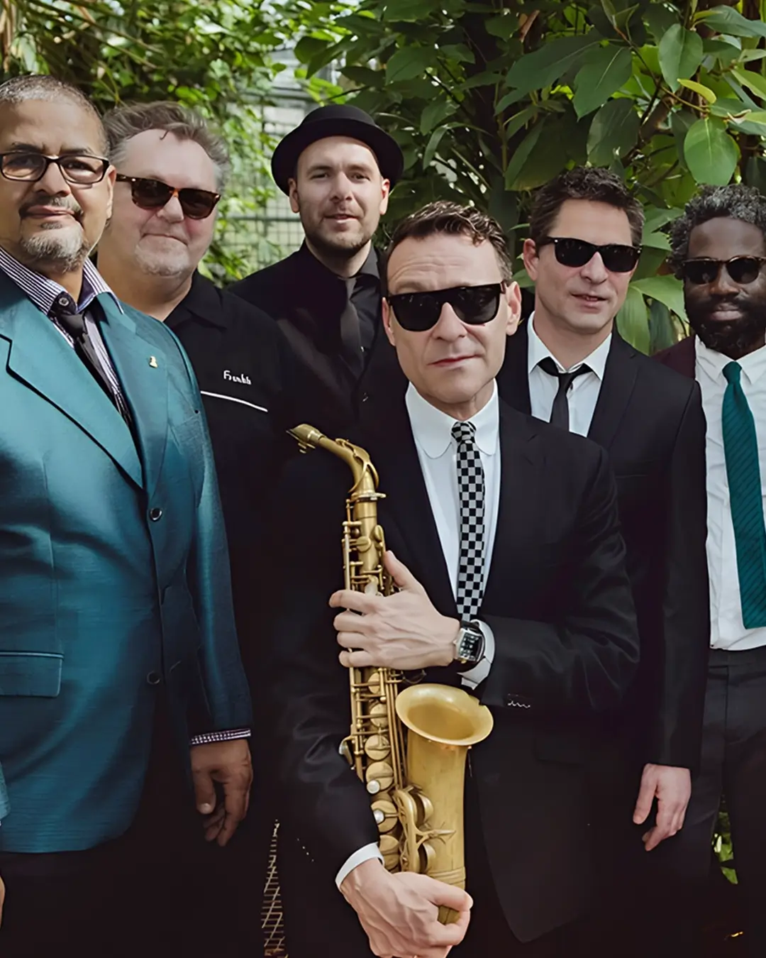 Groepsfoto van een jazzband in een groene, tropische omgeving; meerdere mannen in nette kleding en zonnebrillen poseren samen, met een saxofonist prominent vooraan die zijn instrument vasthoudt.