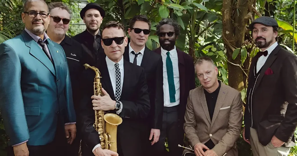 Groepsfoto van een jazzband in een groene, tropische omgeving; meerdere mannen in nette kleding en zonnebrillen poseren samen, met een saxofonist prominent vooraan die zijn instrument vasthoudt.