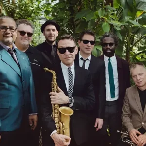 Groepsfoto van een jazzband in een groene, tropische omgeving; meerdere mannen in nette kleding en zonnebrillen poseren samen, met een saxofonist prominent vooraan die zijn instrument vasthoudt.