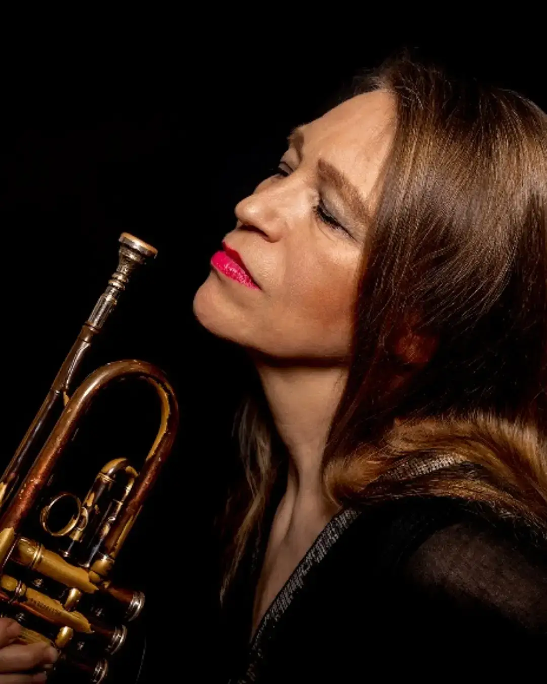 Close-up portret van een vrouwelijke jazztrompettist met roodbruine haren en rode lippenstift, die haar trompet dicht bij haar gezicht houdt en met gesloten ogen lijkt op te gaan in de muziek tegen een donkere achtergrond.