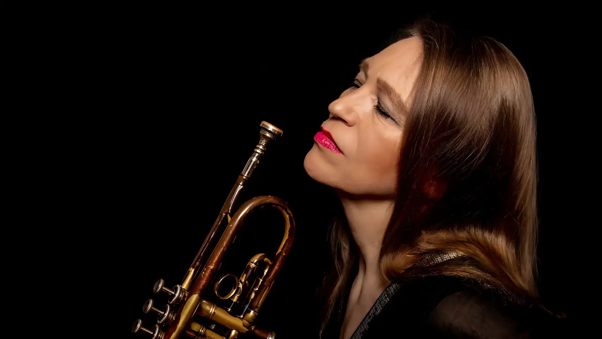 Close-up portret van een vrouwelijke jazztrompettist met roodbruine haren en rode lippenstift, die haar trompet dicht bij haar gezicht houdt en met gesloten ogen lijkt op te gaan in de muziek tegen een donkere achtergrond.