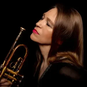 Close-up portret van een vrouwelijke jazztrompettist met roodbruine haren en rode lippenstift, die haar trompet dicht bij haar gezicht houdt en met gesloten ogen lijkt op te gaan in de muziek tegen een donkere achtergrond.