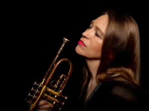 Close-up portret van een vrouwelijke jazztrompettist met roodbruine haren en rode lippenstift, die haar trompet dicht bij haar gezicht houdt en met gesloten ogen lijkt op te gaan in de muziek tegen een donkere achtergrond.