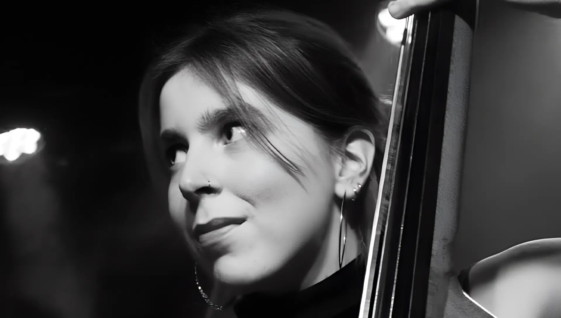 Zwart-wit portret van Veerle van der Wal - contrabassist die met een zachte glimlach opzij kijkt, terwijl ze haar instrument vasthoudt; haar gezicht is half verlicht tegen een donkere achtergrond.