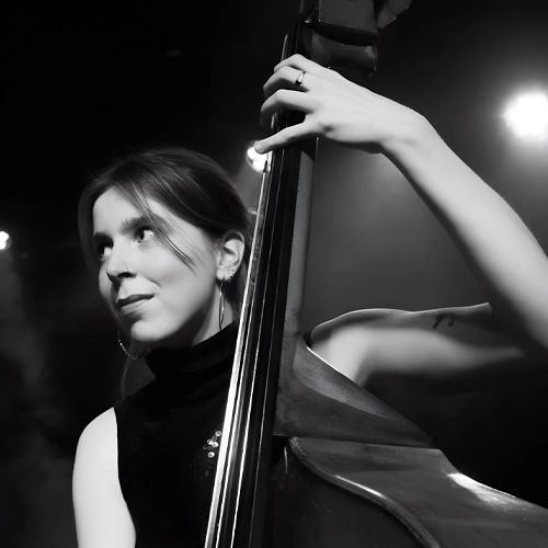 Zwart-wit portret van Veerle van der Wal - contrabassist die met een zachte glimlach opzij kijkt, terwijl ze haar instrument vasthoudt; haar gezicht is half verlicht tegen een donkere achtergrond.