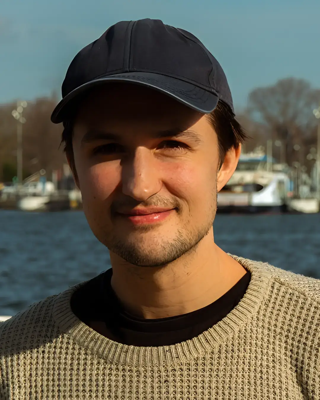 Portret van een jonge man met een zwarte pet en lichte trui, glimlachend aan het water met boten en bomen op de achtergrond in daglicht.
