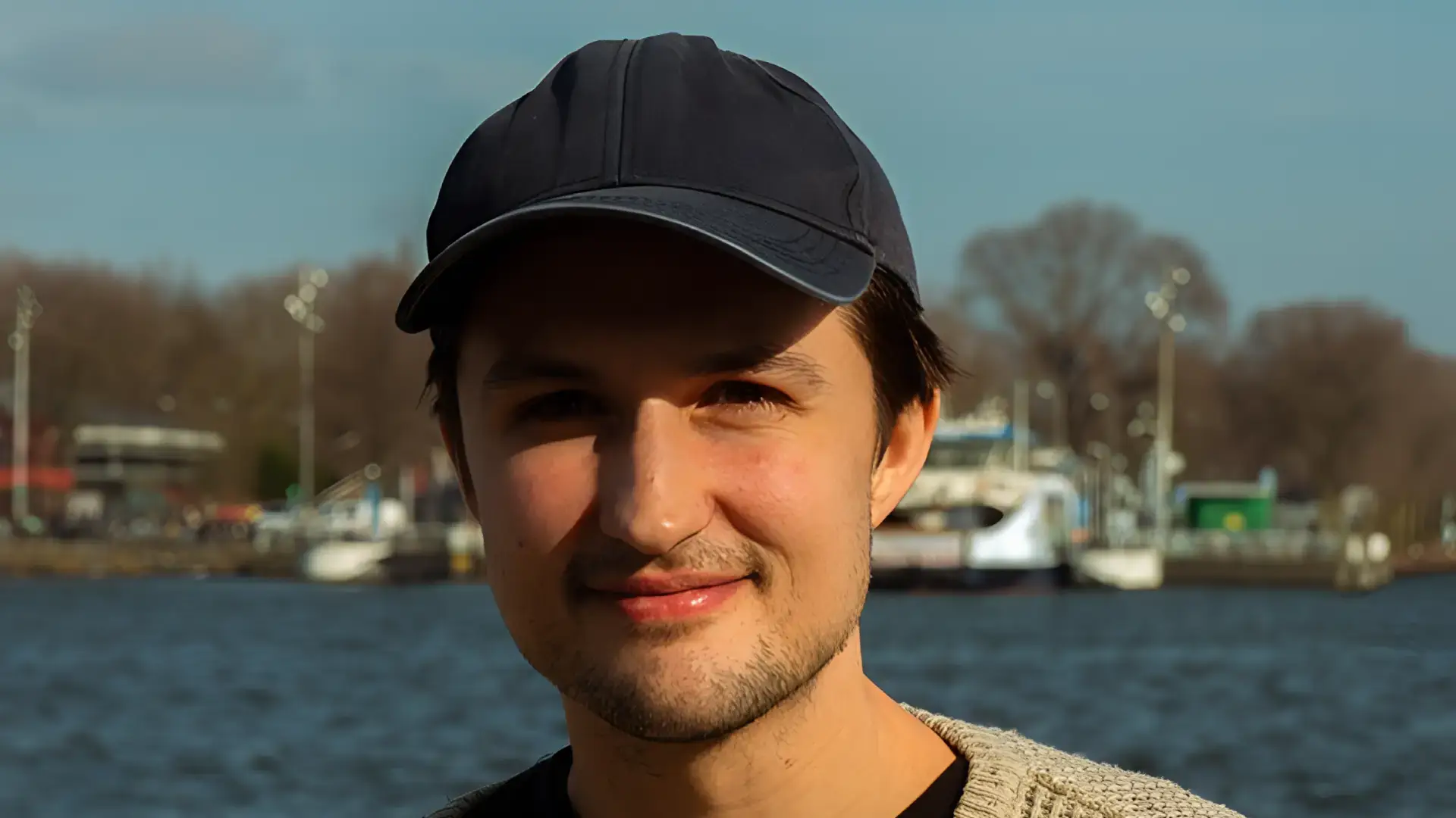 Portret van een jonge man met een zwarte pet en lichte trui, glimlachend aan het water met boten en bomen op de achtergrond in daglicht.