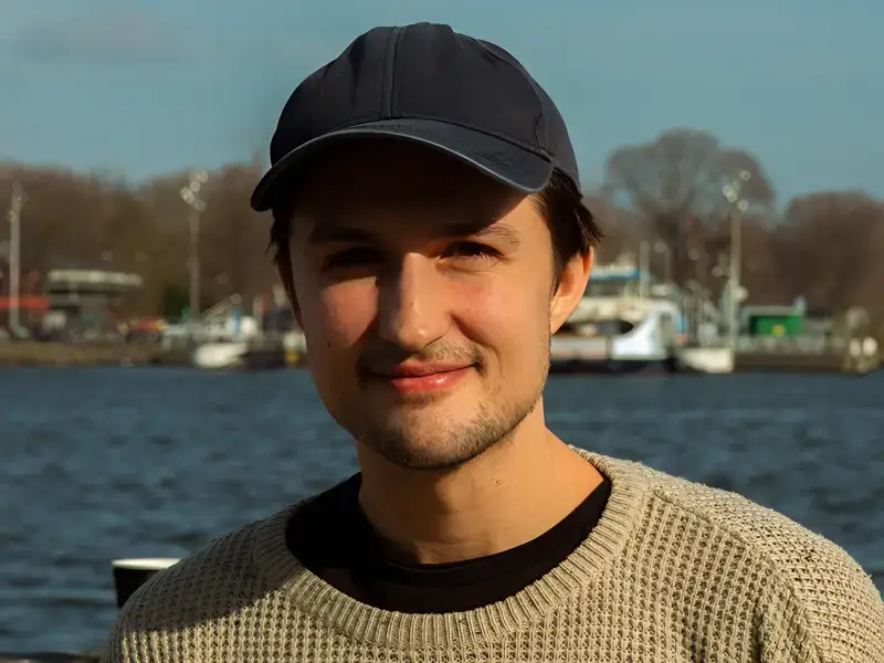 Portret van een jonge man met een zwarte pet en lichte trui, glimlachend aan het water met boten en bomen op de achtergrond in daglicht.