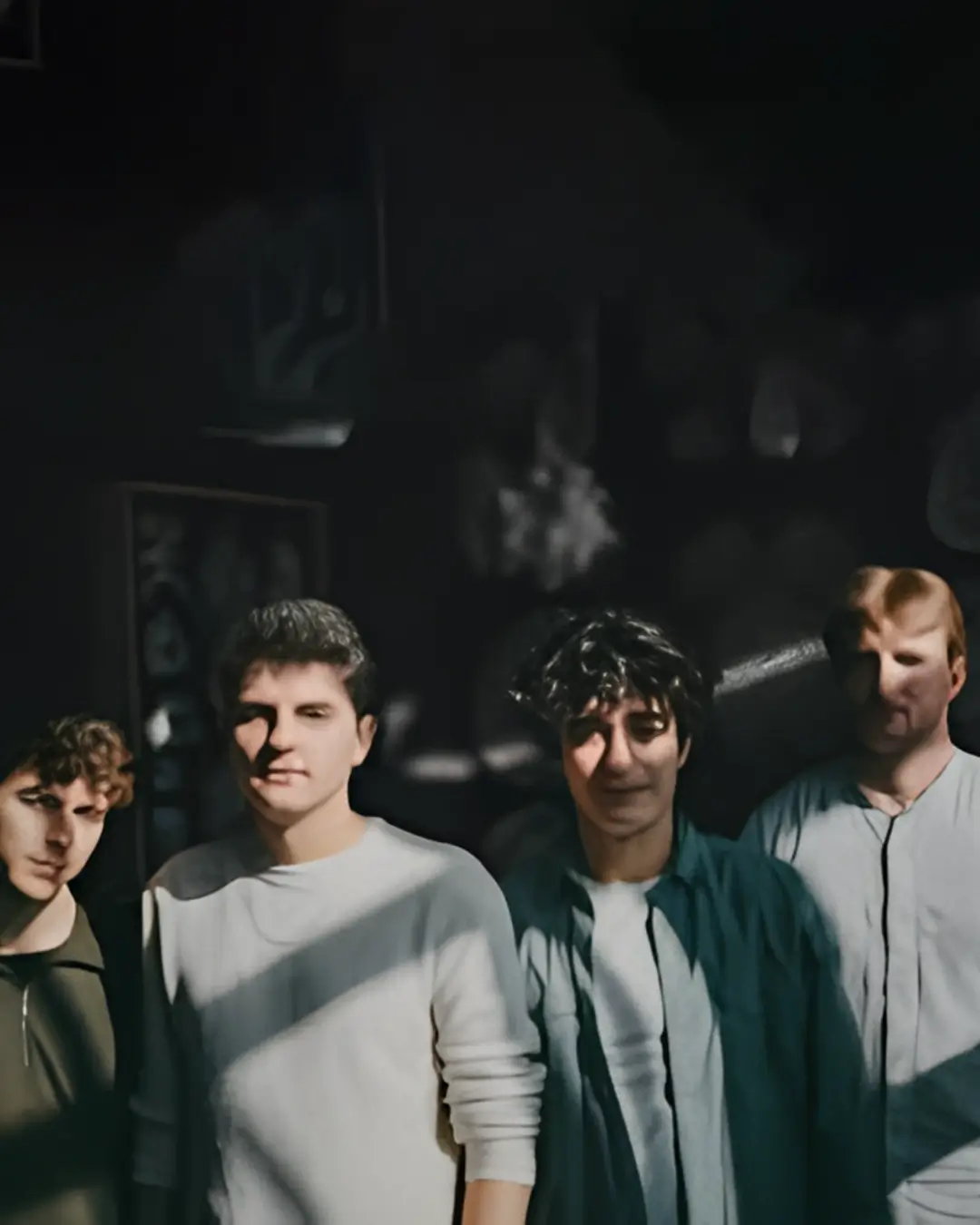 Vier jonge muzikanten staan samen in half schaduw en zonlicht, kijkend naar de camera, met een donkere achtergrond en sterke contrasten.