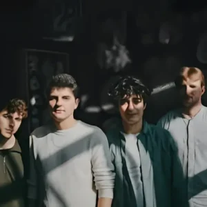 Vier jonge muzikanten staan samen in half schaduw en zonlicht, kijkend naar de camera, met een donkere achtergrond en sterke contrasten.