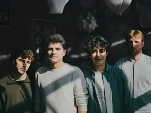 Vier jonge muzikanten staan samen in half schaduw en zonlicht, kijkend naar de camera, met een donkere achtergrond en sterke contrasten.