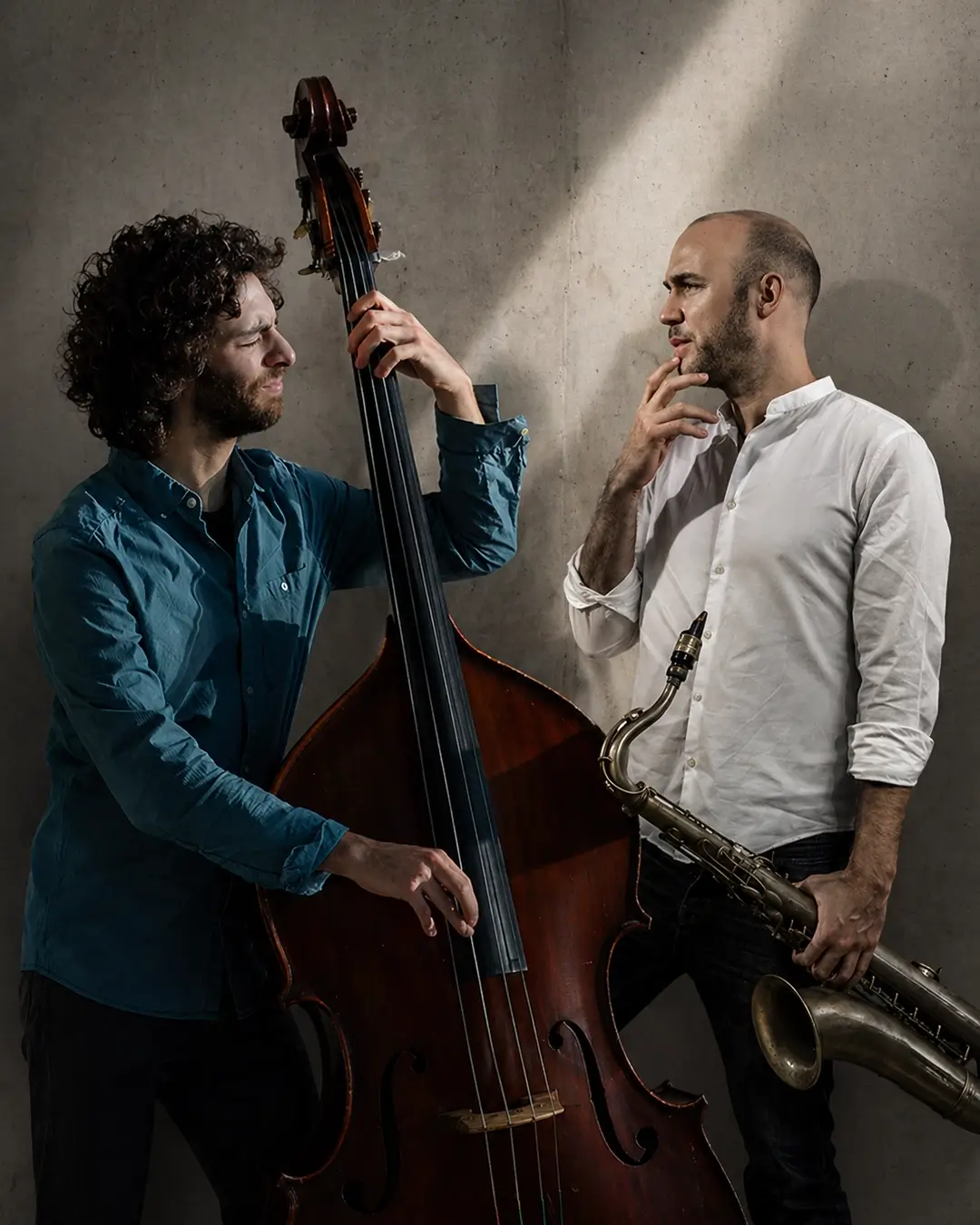 Adán Mizrahi speelt contrabas naast saxofonist Bernard van Rossum, die zijn saxofoon vasthoudt, in een sfeervolle studio-setting met dramatisch licht.