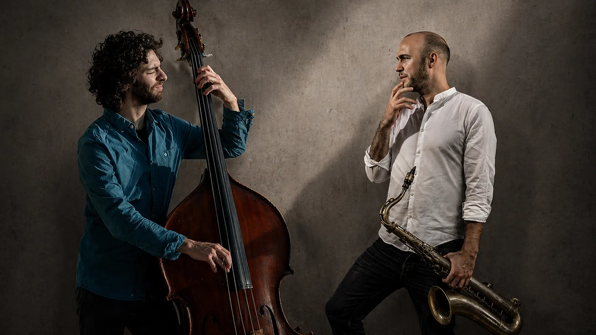Adán Mizrahi speelt contrabas naast saxofonist Bernard van Rossum, die zijn saxofoon vasthoudt, in een sfeervolle studio-setting met dramatisch licht.