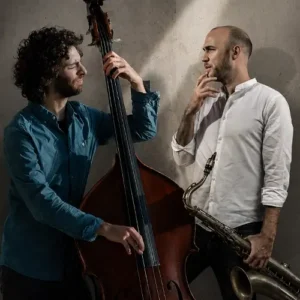 Adán Mizrahi speelt contrabas naast saxofonist Bernard van Rossum, die zijn saxofoon vasthoudt, in een sfeervolle studio-setting met dramatisch licht.