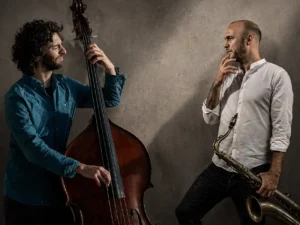 Adán Mizrahi speelt contrabas naast saxofonist Bernard van Rossum, die zijn saxofoon vasthoudt, in een sfeervolle studio-setting met dramatisch licht.