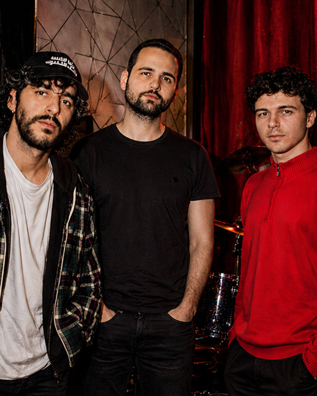Drie bandleden van Mimosa Púdica poseren samen in een donkere ruimte; één draagt een pet, de ander een zwart shirt en de derde een rode trui.
