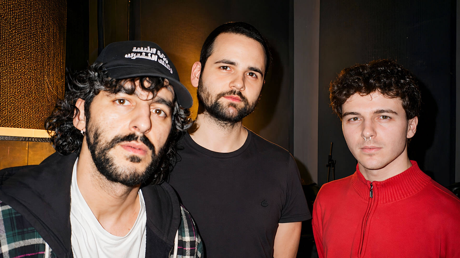 Drie bandleden van Mimosa Púdica poseren samen in een donkere ruimte; één draagt een pet, de ander een zwart shirt en de derde een rode trui.