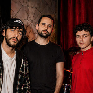 Drie bandleden van Mimosa Púdica poseren samen in een donkere ruimte; één draagt een pet, de ander een zwart shirt en de derde een rode trui.