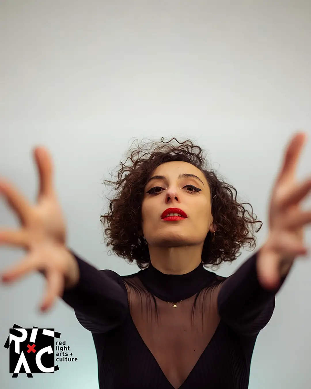 Zangeres en celliste Sanem Kalfa strekt haar armen naar voren tijdens een expressieve performance, gekleed in zwart met rode lippen tegen een lichte achtergrond.