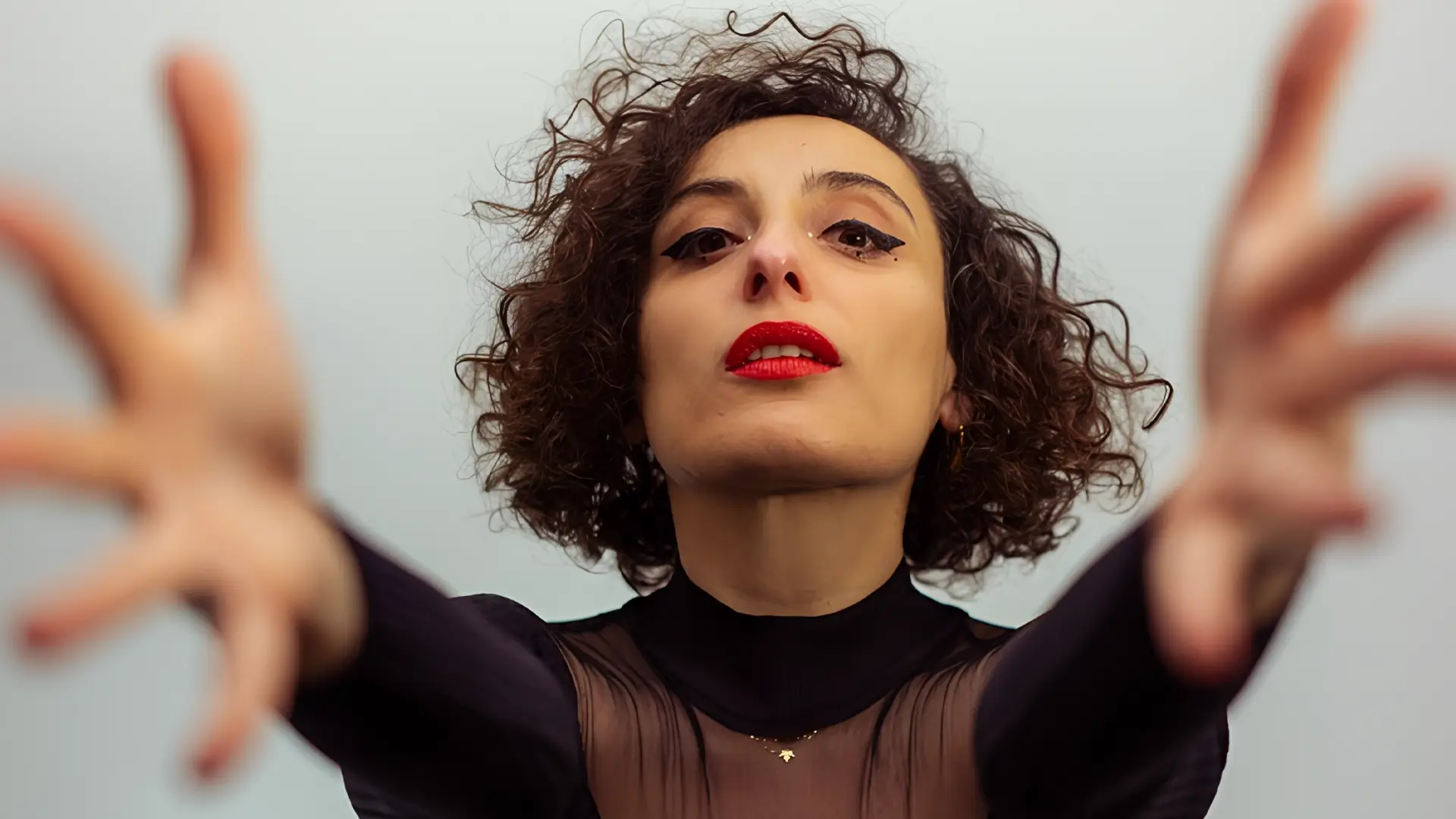 Zangeres en celliste Sanem Kalfa strekt haar armen naar voren tijdens een expressieve performance, gekleed in zwart met rode lippen tegen een lichte achtergrond.