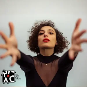 Zangeres en celliste Sanem Kalfa strekt haar armen naar voren tijdens een expressieve performance, gekleed in zwart met rode lippen tegen een lichte achtergrond.