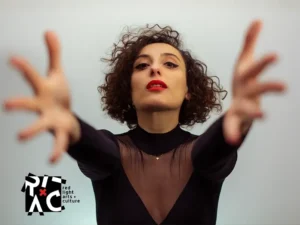 Zangeres en celliste Sanem Kalfa strekt haar armen naar voren tijdens een expressieve performance, gekleed in zwart met rode lippen tegen een lichte achtergrond.