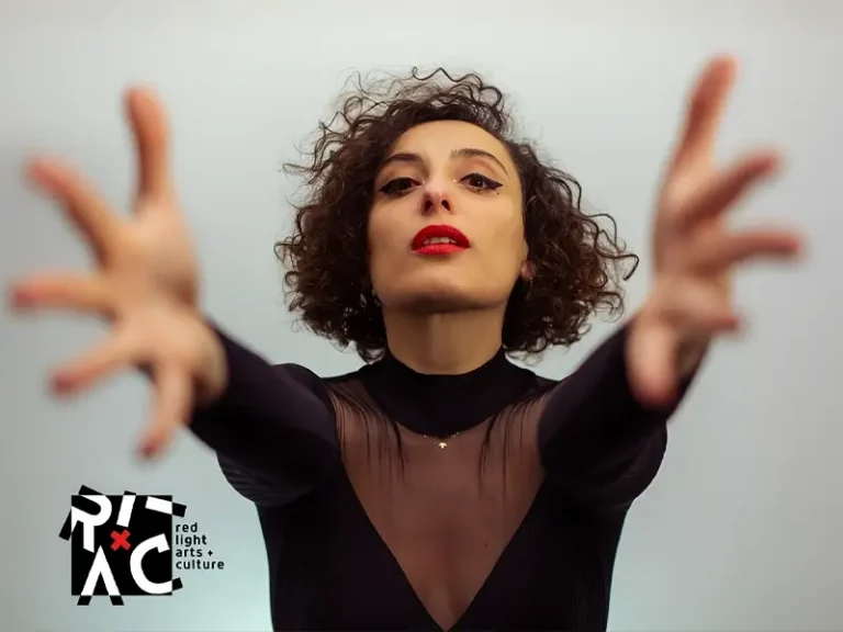 Zangeres en celliste Sanem Kalfa strekt haar armen naar voren tijdens een expressieve performance, gekleed in zwart met rode lippen tegen een lichte achtergrond.