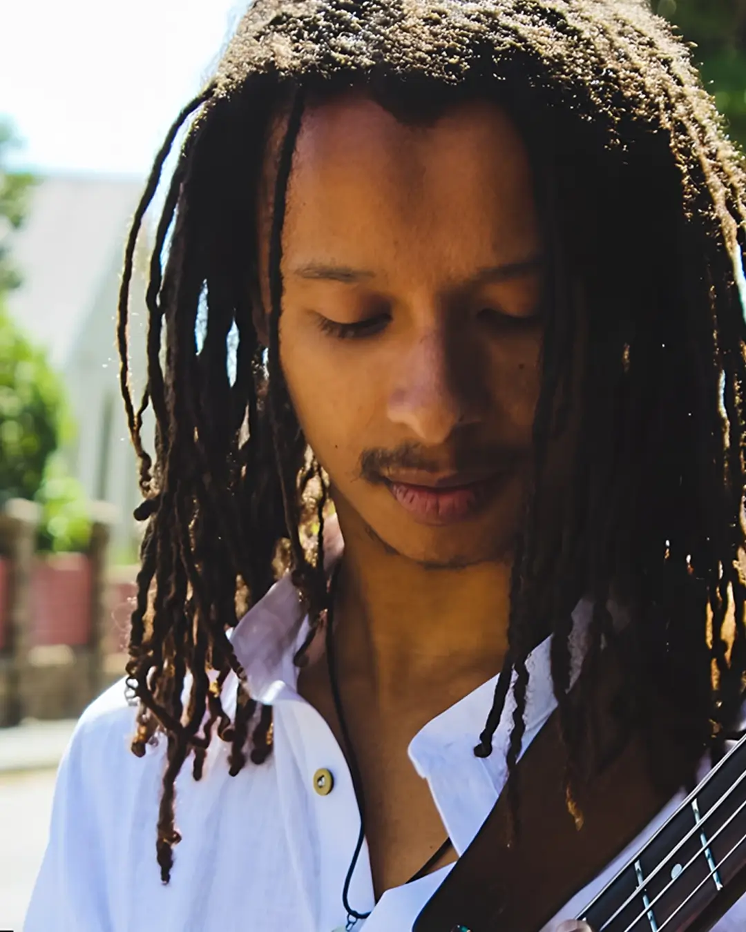 Bassist Zelzele met lange dreadlocks kijkt geconcentreerd naar zijn instrument terwijl hij buiten in het zonlicht speelt.