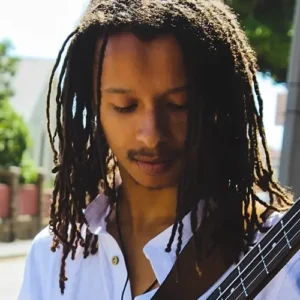 Bassist Zelzele met lange dreadlocks kijkt geconcentreerd naar zijn instrument terwijl hij buiten in het zonlicht speelt.