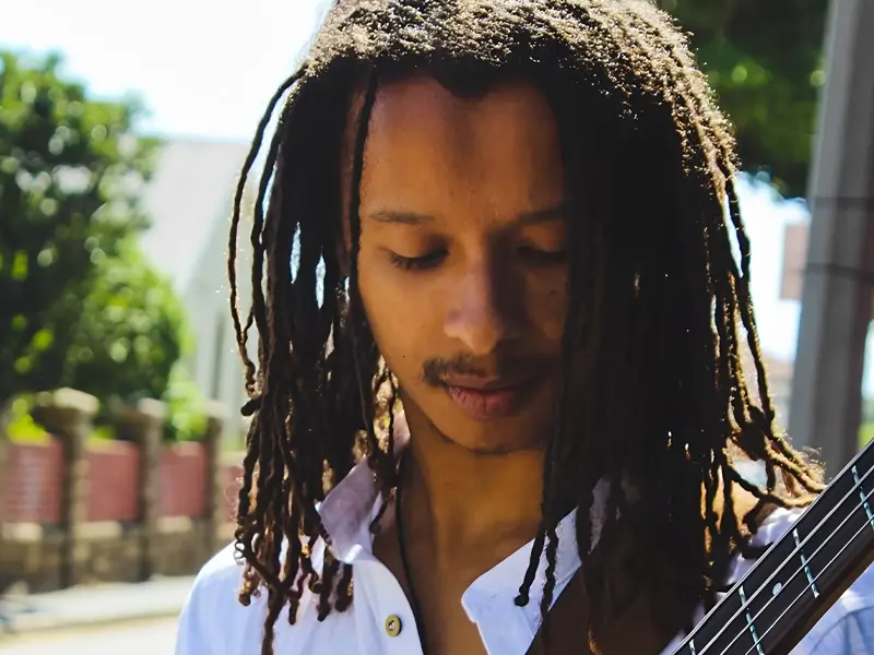 Bassist Zelzele met lange dreadlocks kijkt geconcentreerd naar zijn instrument terwijl hij buiten in het zonlicht speelt.